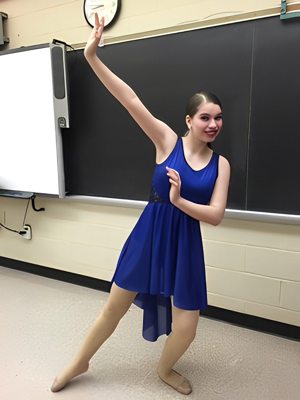 A young dancer in a blue dress poses gracefully in a classroom.